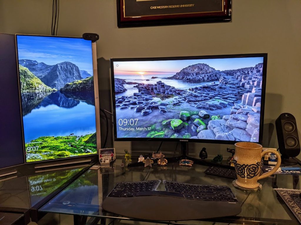 A glass desktop with a keyboard, two monitors, and a ceramic mug. 